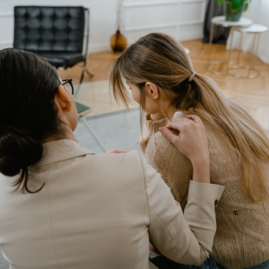 Two individuals seated closely in an office or room. One, wearing a light blazer, gently places a hand on the shoulder of the other, who has long blonde hair in a ponytail and wears a beige knitted sweater.